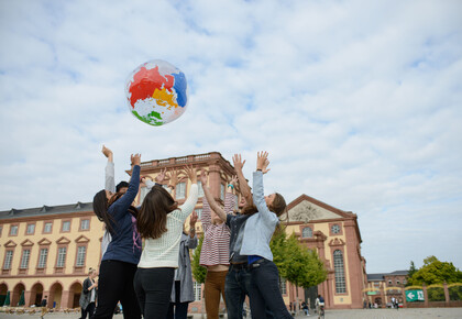 Eine Gruppe von Studierenden wirft eine Weltkugel in die Luft. Im Hintergrund ist das Schloss.