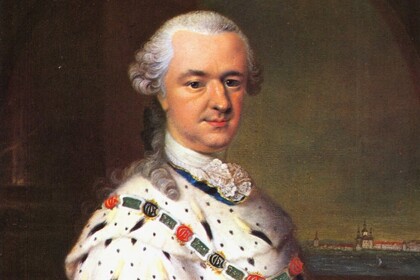 Kurfürst Karl Theodor in rotem Samtgewand mit weißem Hermelinmantel und Ordenskette, hält einen Stab, Hintergrund mit Stadt und Fluss