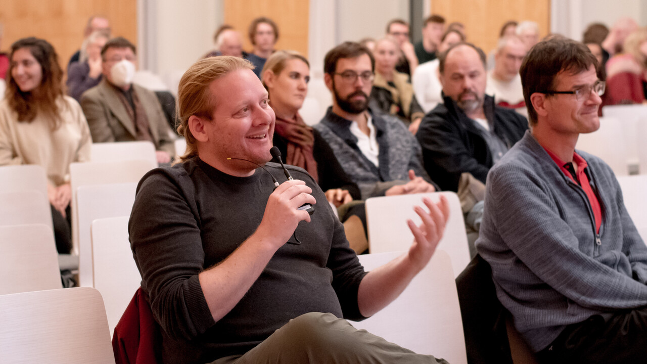 Frage aus dem Publikum: Zentral im Bild hält ein Mann in schwarzem Pollover und beiger Hose ein Funkmikrofon in seiner rechten Hand in Mundhöhe und spricht lächelnd hinein. Mit seiner linken Hand gestikuliert er mit sichtbarer Bewegungsunschärfe im Bild vor sich. Die Kamera ist schräg links vor ihm platziert, um ihn herum sitzen weitere herbstlich gekleidete Menschen bei einigen freien Plätzen auf Stühlen. Im hinteren, leicht unscharfen Bildbereich fällt ein einzelner Mann mit einer Atemschutzmaske auf. Den unscharfen Hintergrund bildet die abwechselnd weiß und holzfarbene Fensterfront der Universitätsaula.