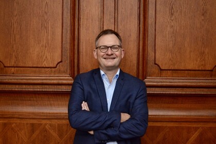 Philipp Gassert vor dem Eingangsportal im Ehrenhof der Universität Mannheim. Er trägt Hemd und Anzugjacke, seine Arme sind vor der Brust verschränkt. Er trägt eine Brille und lächelt in die Kamera.  