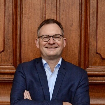 Philipp Gassert vor dem Eingangsportal im Ehrenhof der Universität Mannheim. Er trägt Hemd und Anzugjacke, seine Arme sind vor der Brust verschränkt. Er trägt eine Brille und lächelt in die Kamera.  