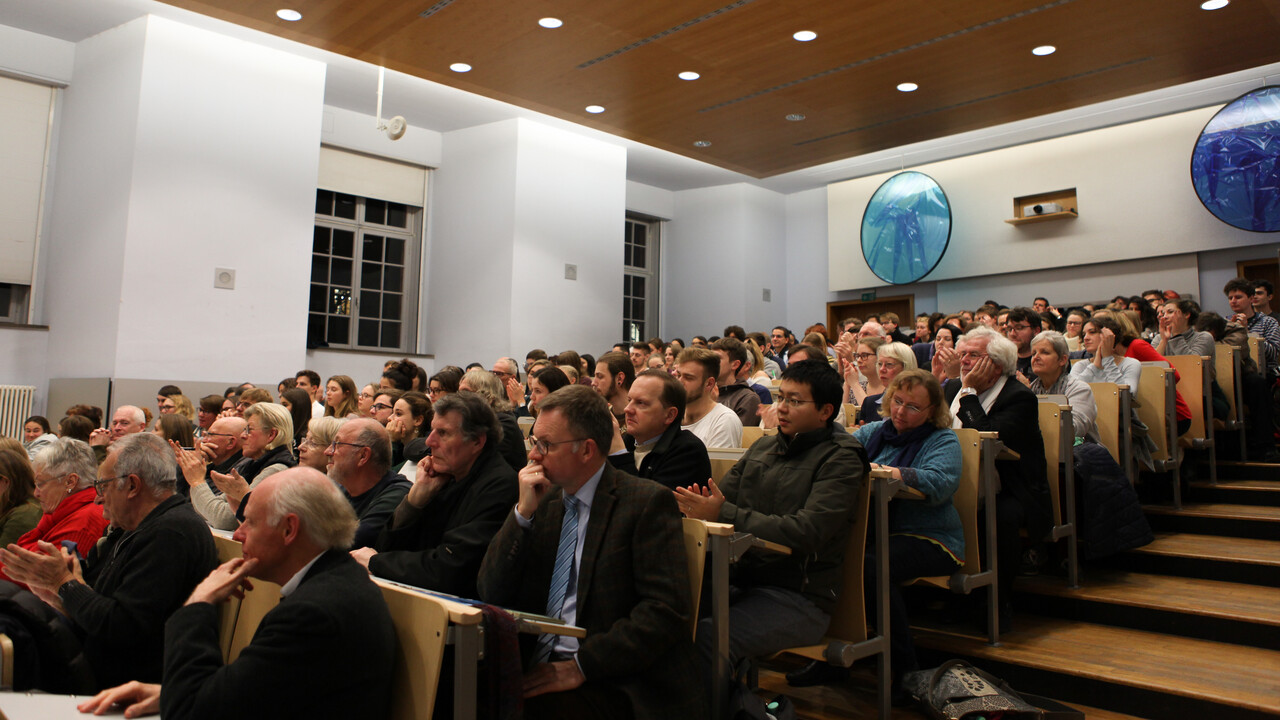 Blick von schräg unten auf das Publikum, das auf den zum Vortragspult abfallenden vollbesetzten Sitzplatzreihen des Hörsaals Platz genommen hat. In der oberen Bildhälfte ist eine weiße Wand mit Fenstern, rechts, eine Holzdecke mit Lichtstrahlern sowie die Rückwand des Saals mit Türen und Multimediaprojektor zu sehen. Am rechten Bildende ist ein treppenartiger Aufgang angeschnitten.