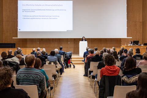 Welttag der Philosophie 2025: Blick aus dem Mittelgang zwischen dem Stuhlreihen mit Publikum auf die Bühne beim Vortrag von Prof. Dr. phil. Elif Özmen in der Aula der Universität Mannheim mit ihrer Holzvertäfelung bei abgesenkter Leinwand mit darauf projizierter Vortragspräsentation.