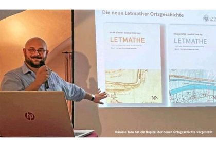 Zeitungsausschnitt mit einem männlichen Vortragenden vor der Leinwand, die ein Buchcover zeigt