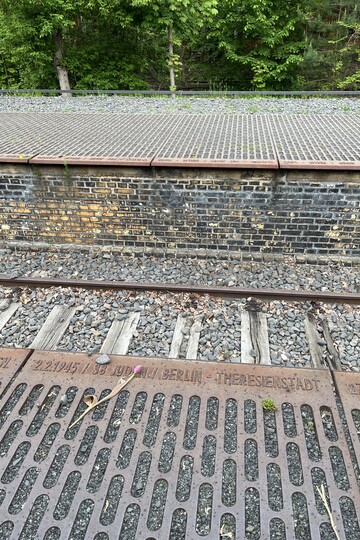 Mahnmal Gleis 17: Blick ins Gleisbett. Am Rand des Bahnsteigs stehen die Deportationstransporte mit Datum, Personenzahl und Destination. Davor liegt eine Blume. 