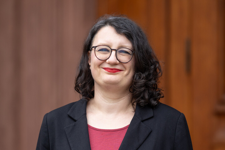 A middle-aged woman stands in front of a blurred, brown-orange background (presumably a wooden wall or door). She has fair skin and shoulder-length, wavy, dark brown hair with a side parting. She wears round, dark-framed glasses, red lipstick, and a friendly smile. She is wearing a black blazer over a burgundy top.