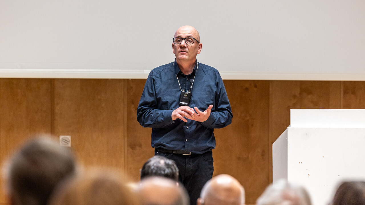 Begrüßung durch Prof. Dr. Wolfgang Freitag Prof. Dr. Wolfgang Freitag mit umgehängtem Funkmikrofon auf der Bühne neben dem Rednerpult stehend als Eröffnung der Veranstaltung zum Tag der Philosophie am 20.11.2025. Er trägt dunkle Kleidung und eine Brille. Im Hintergrund Holzvertäfelung und Leinwand mit angeschnittener Projektion, im Vordergrund unscharf Hinterköpfe des sitzendes Saalpublikums.