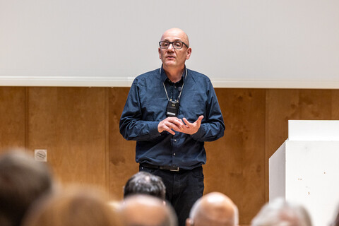 Prof. Dr. Wolfgang Freitag mit umgehängtem Funkmikrofon auf der Bühne neben dem Rednerpult stehend als Eröffnung der Veranstaltung zum Tag der Philosophie am 20.11.2025. Er trägt dunkle Kleidung und eine Brille. Im Hintergrund Holzvertäfelung und Leinwand mit angeschnittener Projektion, im Vordergrund unscharf Hinterköpfe des sitzendes Saalpublikums.