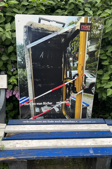 Eine plakatierte Holzplatte, die aufrecht auf einer weiß-blau gestreiften Holzbank steht. Darauf abgebildet ist das Foto des Bücherschrankes nach dem Brandanschlag 2023, das Innere ist ausgebrannt und verkohlt. Darunter steht in weißen Lettern das Zitat: "Dort, wo man Bücher verbrennt, verbrennt man am Ende auch Menschen" (Heinrich Heine) 