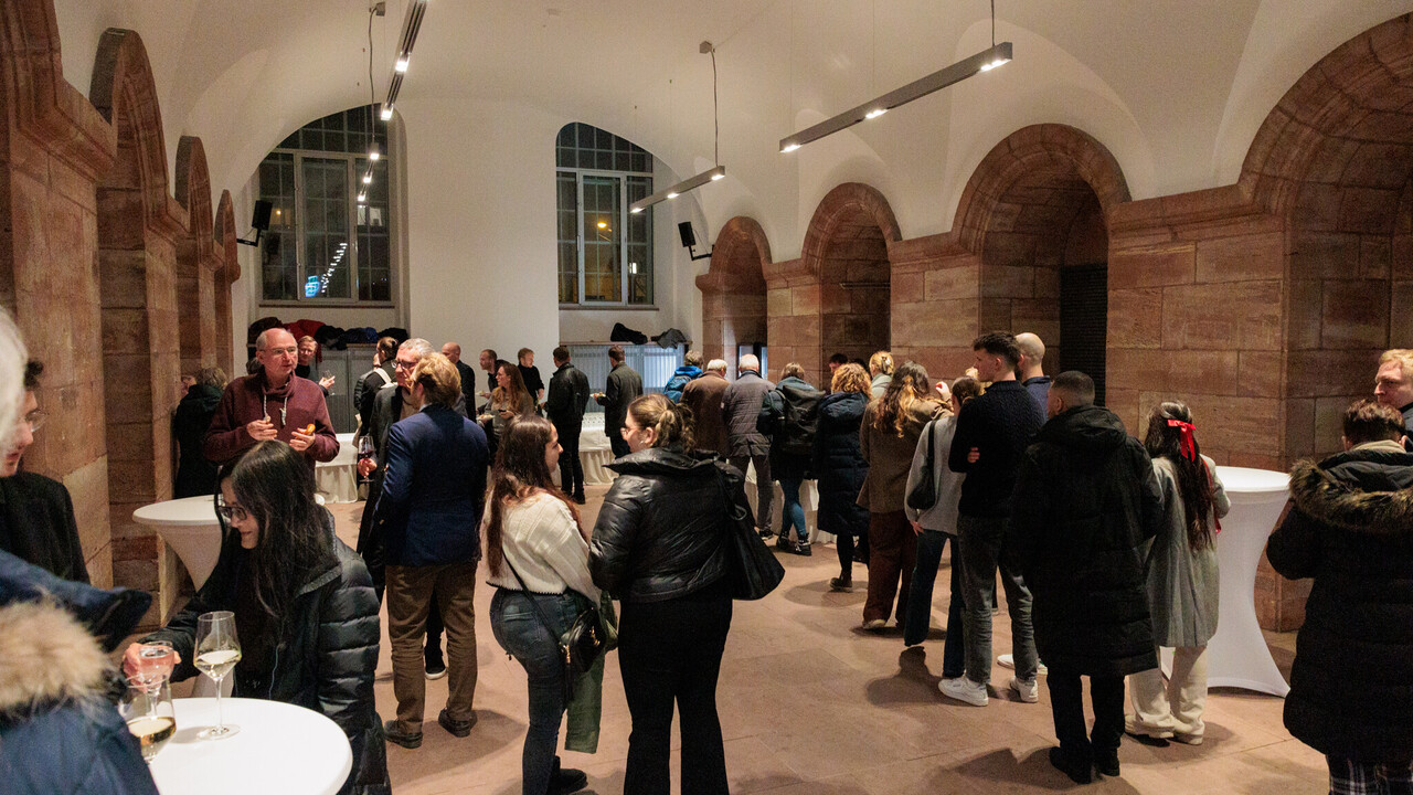 Impressionen vom Empfang in den „Katakomben“ Empfang in den Katakomben der Universität Mannheim am Tag der Philosophie am 21.11.2024: Totale eines Bereichs zwischen 2 Säulengängen mit Rundbögen aus rotem Sandstein und weißer Wand sowie Decke mit etwa 2 Dutzend Personen im Gespräch und am Buffet.