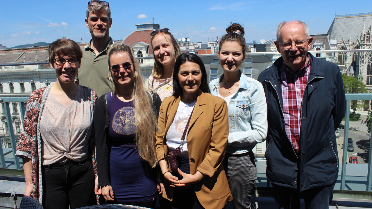 Gruppenfoto von 7 Personen in Freizeitkleidung vor einem Geländer. Im Hintergrund sind Fassaden und Dächer zu sehen sowie rechts im Anschnitt eine Straße und ein Platz mit grünen Laubbäumen vor einer Kirche.