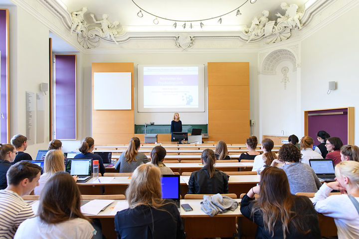 Studierende sitzen in einem Vorlesungssaal. Der Blick ist auf eine Dozentin vor einer PowerPoint Präsentation gerichtet.