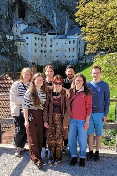 6 Mannheimer Studierende und eine Dozentin stehen vor einer Burg, die vor eine Felsenhöhle gebaut ist, rechts ein Baum und eine grüne Wiese.