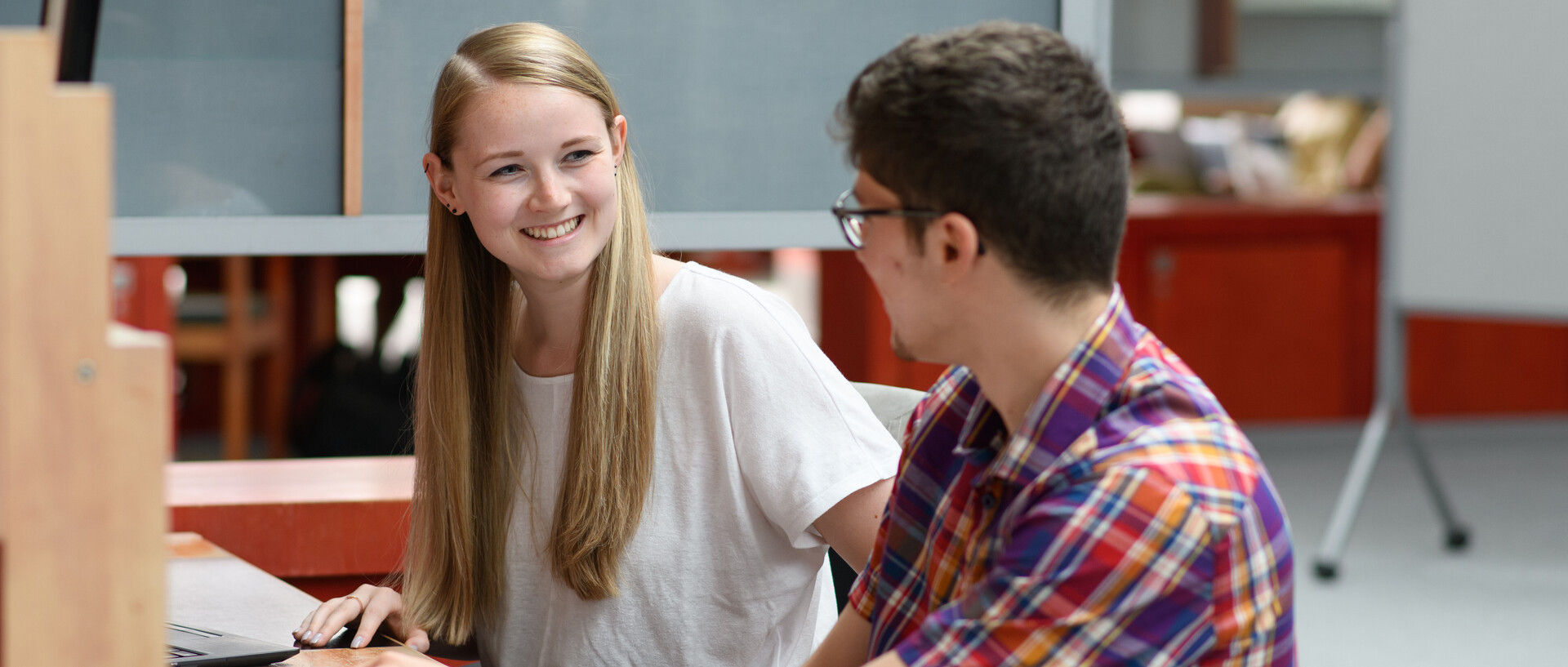 Eine Studentin und ein Student sitzen lachend an einem der Arbeitstische in der A3-Bibliothek.