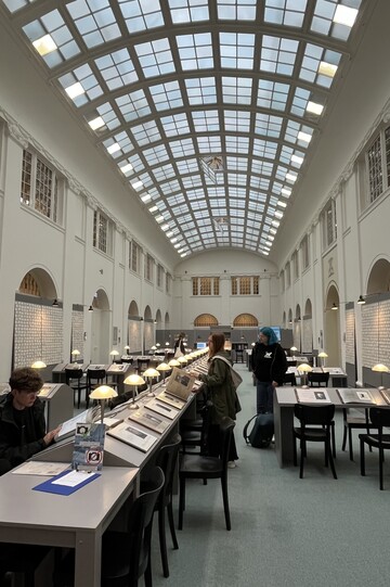 Großer, heller Lesesaal mit gewölbter Glasdecke. Mehrere Menschen sitzen oder stehen an langen Tischen und betrachten historische Dokumente oder Bücher in einer ruhigen Ausstellungs- bzw. Bibliotheksatmosphäre.