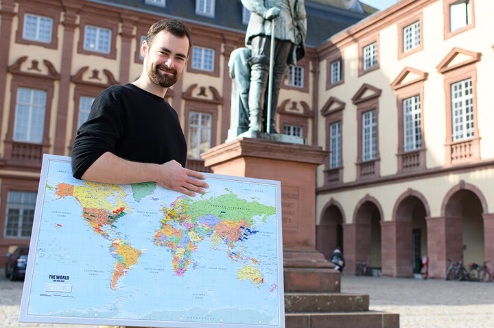 Ein Mann steht auf dem Ehrenhof und trägt eine Weltkarte unterm Arm. Er hat ein schwarzes Langarmshirt an
