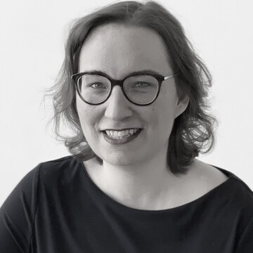 Charlotte Lerg trägt ein dunkles Oberteil, hat schulterlanges Haar und eine Brille. Sie lächelt. 