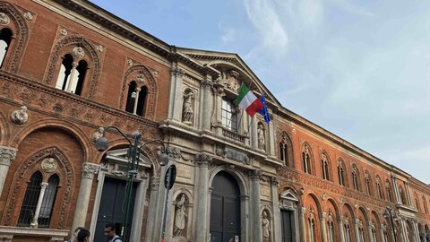 Historisches Universitätsgebäude mit roten Backsteinfassaden, verzierten Fenstern und italienischer sowie EU-Flagge am Eingang