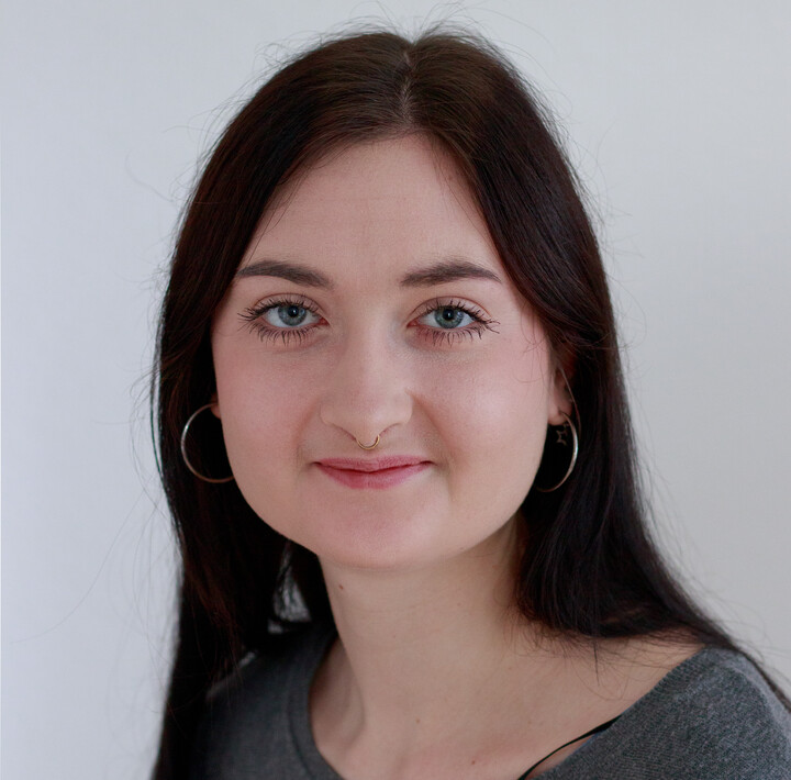 Benita Baum trägt ein graues Oberteil. Sie hat lange dunkle Haare, trägt silberne Ohrringe und lächelt. 
