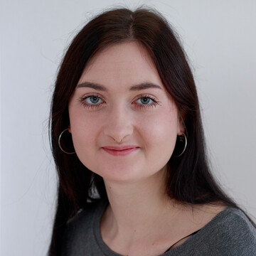 Benita Baum trägt ein graues Oberteil. Sie hat lange dunkle Haare, trägt silberne Ohrringe und lächelt. 