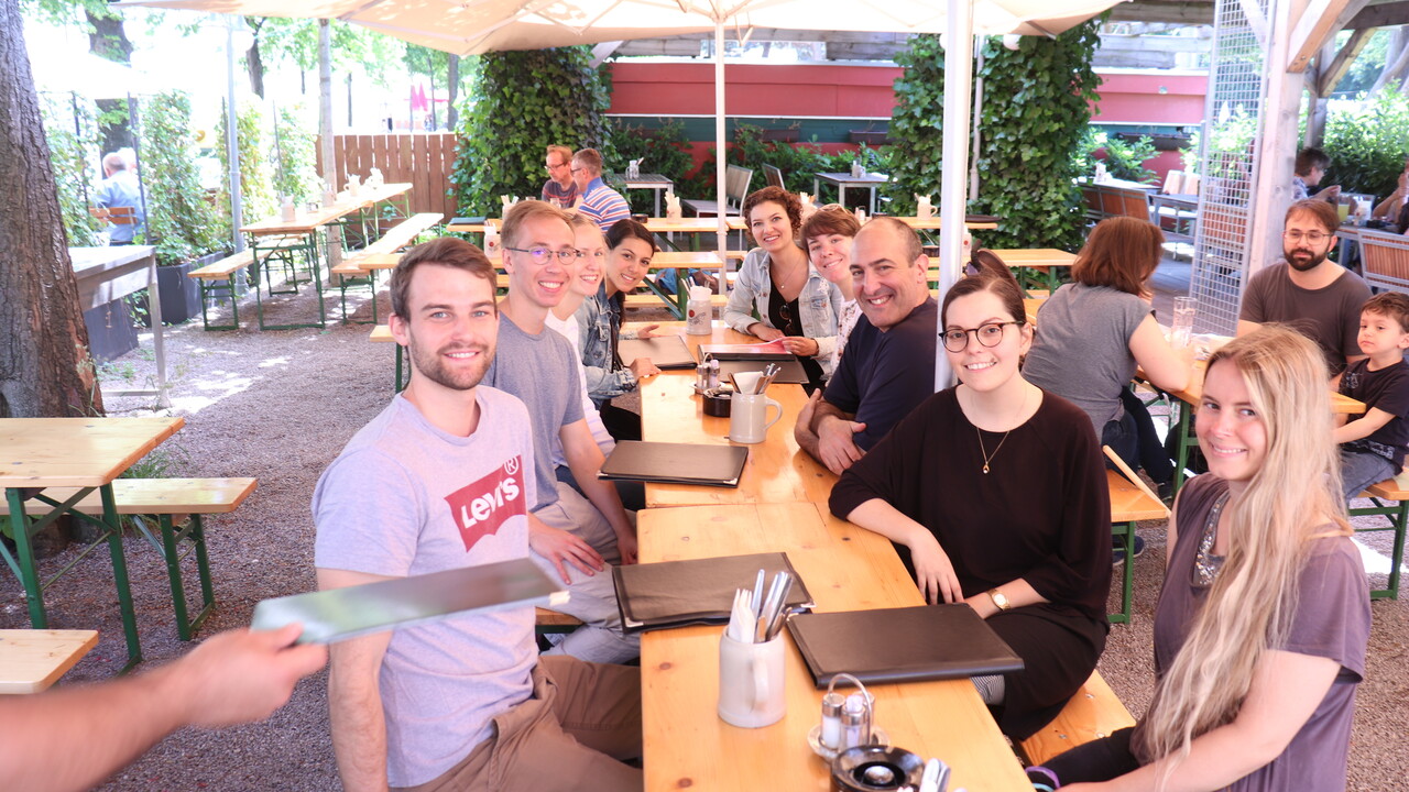 9 Personen in Freizeitkleidung, die offensichtlich eine Gruppe bilden und alle in die Kamera schauen, sitzen an 2 Biergarnituren unter einem großen quadratischen Sonnenschirm in einem sonnigen Außenbereich einer Gaststätte. Auf den Tischen befinden sich Speisekarten, Besteck und Aschenbecher, eine weitere Karte wird von links ins Bild hineingereicht. Im Hintergrund sitzen an einigen Plätzen weitere Personen. Das Ambiente ist durch Bäume, von denen man einen Stamm links im Anschnitt sieht und Efeu an Pergulabalken begrünt.