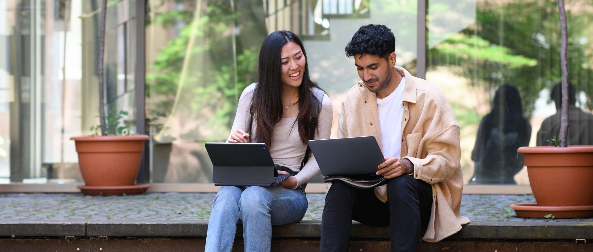 Eine Studentin und ein Student sitzen lachend auf einer Treppe und arbeiten an ihren Laptops. Link: Digitaler Master-Infotag am 27. Februar 2024.