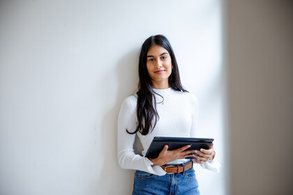Eine junge Frau steht vor einer Wand. Sie hält ein Tablet in der Hand und lächelt in die Kamera.
