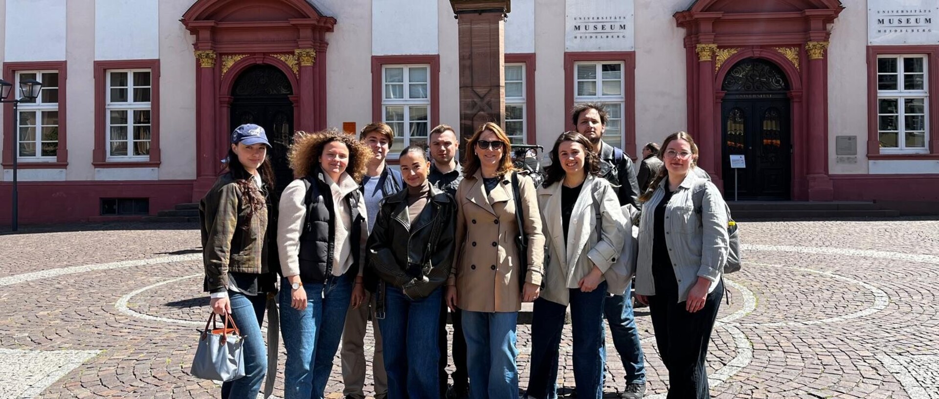 Eine Gruppe von 9 Personen steht vor einem Brunnen auf einem gepflasterten Platz vor einem historischen Gebäude.
