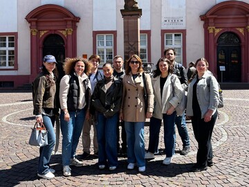 Eine Gruppe von 9 Personen steht vor einem Brunnen auf einem gepflasterten Platz vor einem historischen Gebäude.