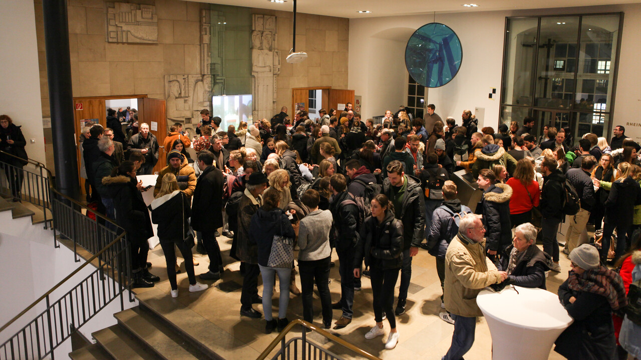 Von schräg oben aus den sich links befindlichen Treppenaufgängen aufgenommene Totale auf mehrere Dutzend Menschen im Foyer der großen Hörsääle im Schloss, die in winterlicher Kleidung in Gruppen, teilweise um Stehtische herum, miteinander im Austausch stehen. Im Hintergrund geöffnete Holztüren zum Hörsaal in künstlerisch gestalteter heller Wand und rechts daran anschließend eine weiße Wand mit großer Glasfront. In der hellen Decke sind 8 eingelassene Beleuchtungselemente zu sehen.