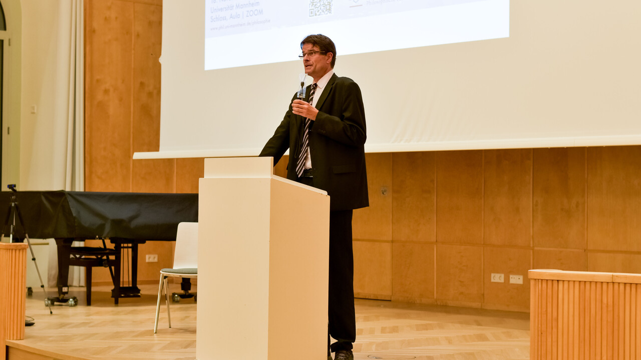 Prof. Dr. Bernward Gesang steht, bekleidet mit schwarzem Anzug, weißem Hemd und in ebenfalls diesen Farben gestreifter Krawatte dunkler Holzvertäfelung und heller Projektionsfläche mit angeschnittener projizierter Powerpoint-Folie hinter einem hellen Pult auf der Bühne. Ein trägt eine Brille und ein umgehängtes Funkmikrofon, das er mit der linken Hand etwas näher vor den Mund hält. Im Hintergrund auf der linken Seite ist außerdem ein Piano und ein Mikrofonständer zu sehen.