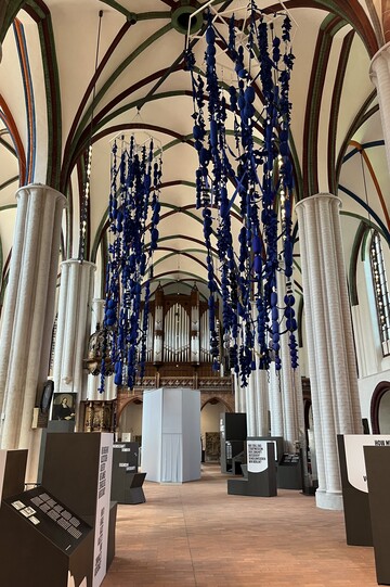 Das Innere der Nikolaikirche in Berlin, fotografiert in Richtung Eingang. Säulen und Decke sind weiß, im Hintergrund befindet sich eine Orgel. Von der Decke hängen blaue Stoffbahnen (eine Kunstinstallation). Im Innenraum befinden sich die Infotafeln einer Ausstellung. 