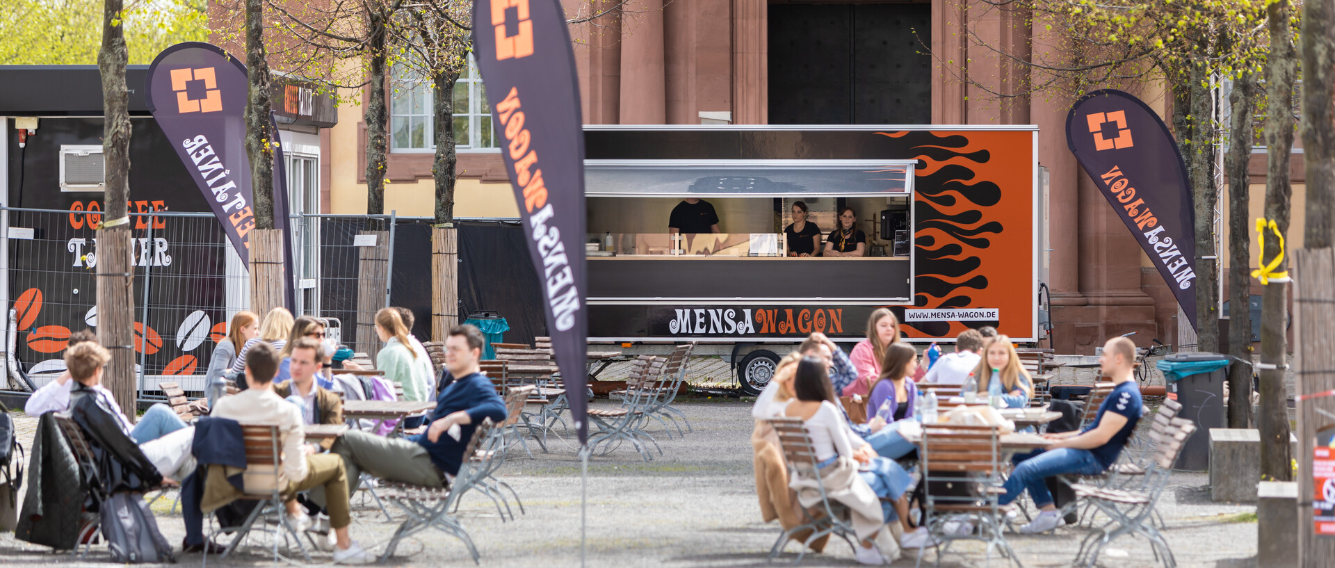 Mensawagon der Uni vor dem Mannheimer Schloss. Vor dem Wagon sitzen Studierende entspannt an Café-Tischen.