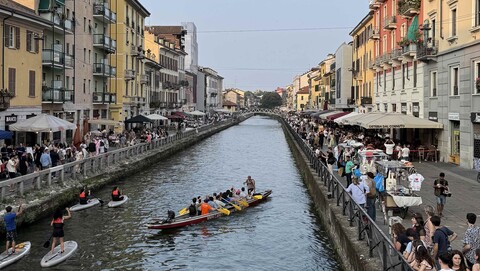 Menschen sitzen und gehen entlang des Kanals Navigli in Mailand, mit Booten auf dem Wasser und mehrstöckigen Gebäuden beidseitig