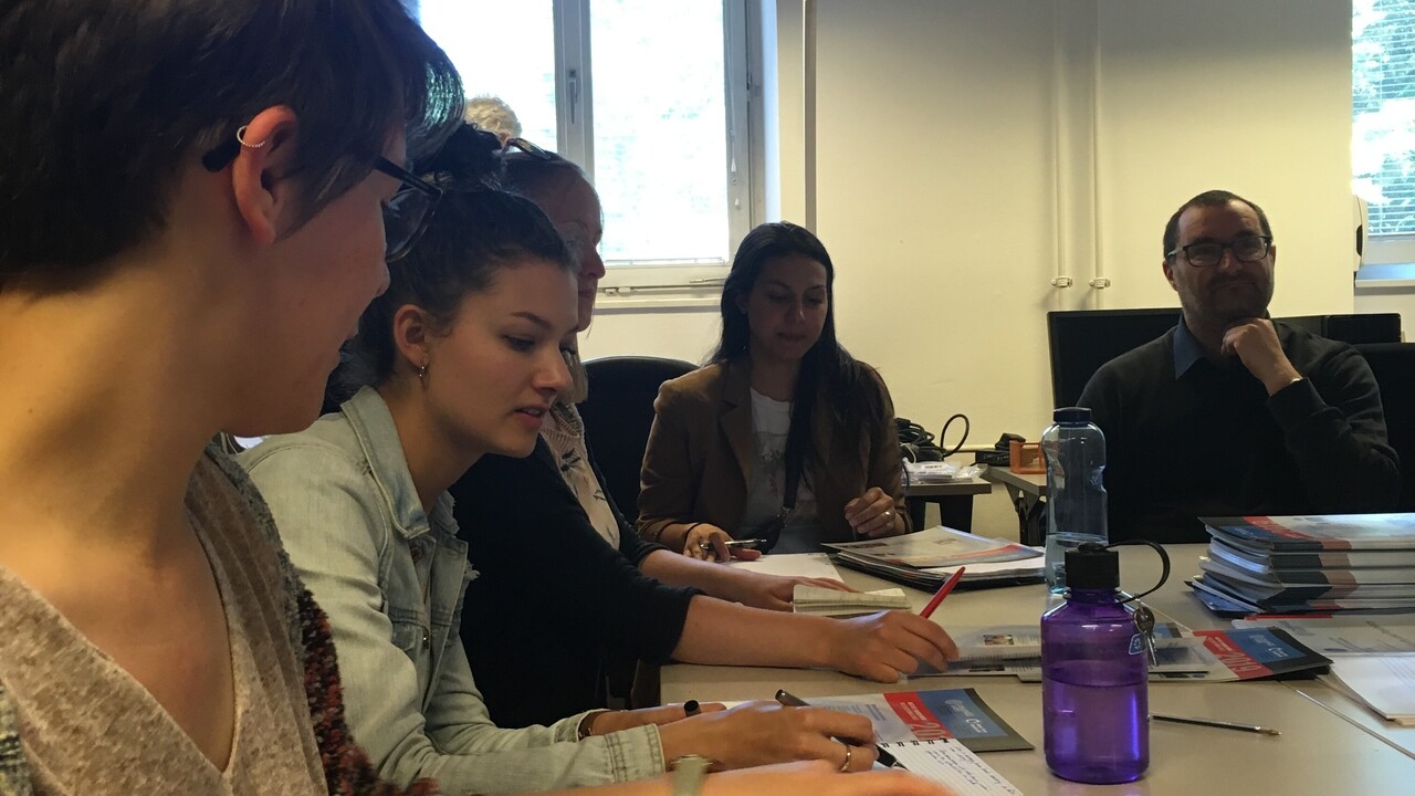 5 Personen sitzend um L-förmig gestellte Tische, auf denen Schreibutensilien, Unterlagen und Getränkeflaschen zu sehen sind.