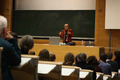 Blick aus dem Publikum auf die Vortragende Prof. Dr. Ursula Wolf. Man sieht die zur dunkelgrünen Tafel und Pult abfallenden Sitzplatzreihen mit den Köpfen des Auditoriums von hinten. Prof. Wolf steht in rot- und schwarztönen herbstlich gekleidet zwischen Pult und Tafel und hebt gerade gestikulierend Unterarme und Hände etwas nach oben.