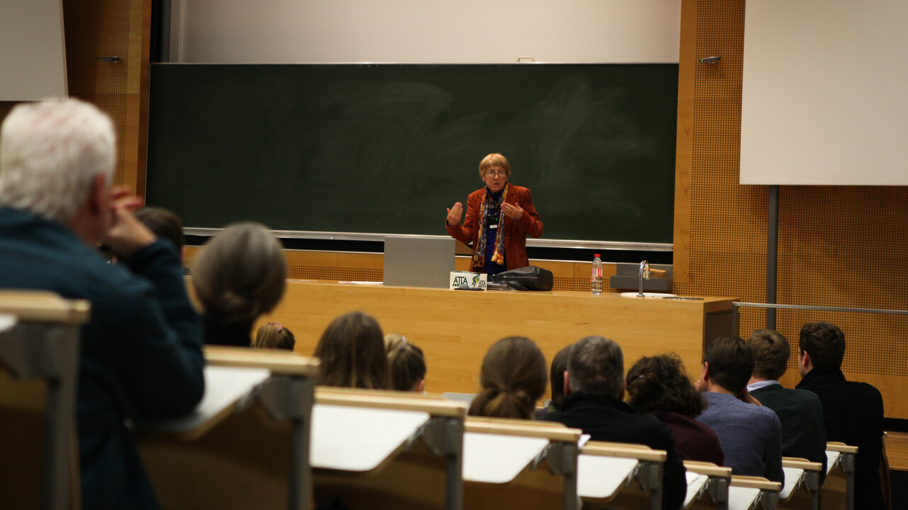 Blick aus dem Publikum auf die Vortragende Prof. Dr. Ursula Wolf. Man sieht die zur dunkelgrünen Tafel und Pult abfallenden Sitzplatzreihen mit den Köpfen des Auditoriums von hinten. Prof. Wolf steht in rot- und schwarztönen herbstlich gekleidet zwischen Pult und Tafel und hebt gerade gestikulierend Unterarme und Hände etwas nach oben.