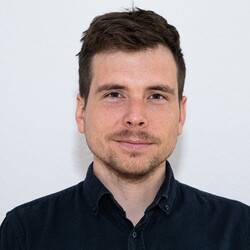 Mann mit kurzen lockigen Haaren, Bart und schwarzem Hemd, vor hellem Hintergrund