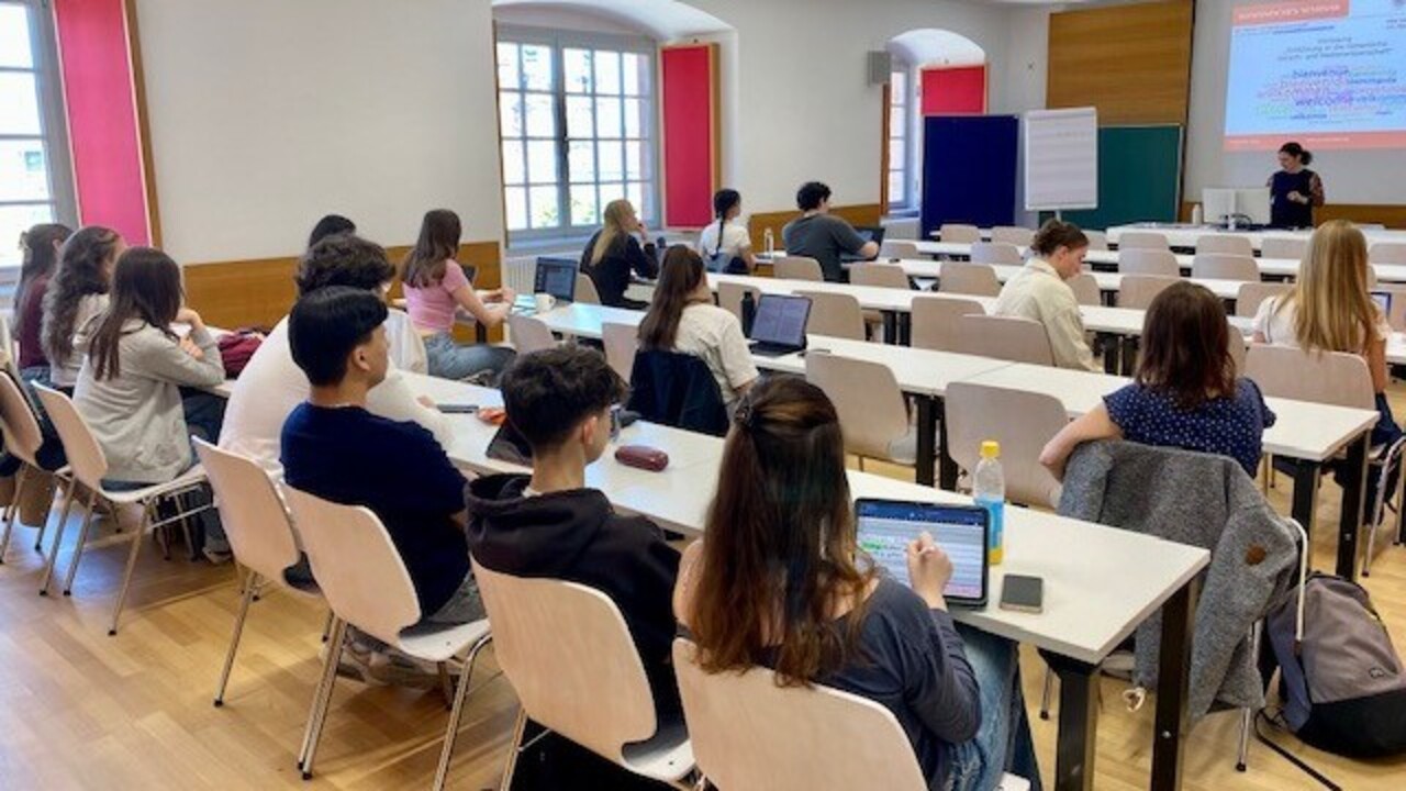 Schüler*innen eines Gymnasiums sitzen in einem Hörsaal. Ihr Blick ist zur Dozentin gerichtet.