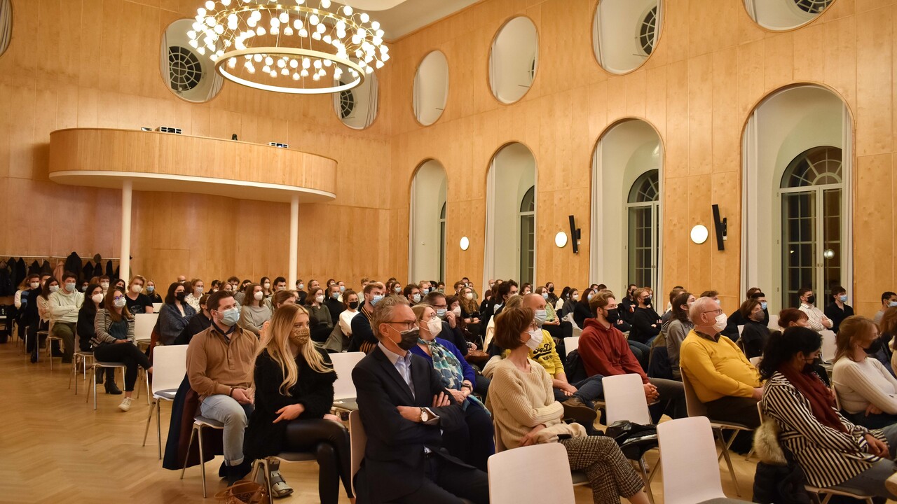 Blick von vorne auf das Publikum in der holzgetäfelten, von rundlichen Fensteröffnungen und mit aufwendigen Deckenlampen geprägten Aula der Universität Mannheim. Die herbstlich gekleideten Menschen sitzen mit Corona-Atemschutzmasken auf leicht auseinandergezogenen Stuhlreihen, von denen die meisten besetzt sind.