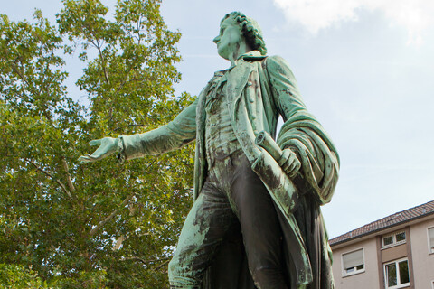 Eine Statue von Friedrich Schiller aus Metall. Die Figur ist grün verfärbt und steht auf grauen Steinen.