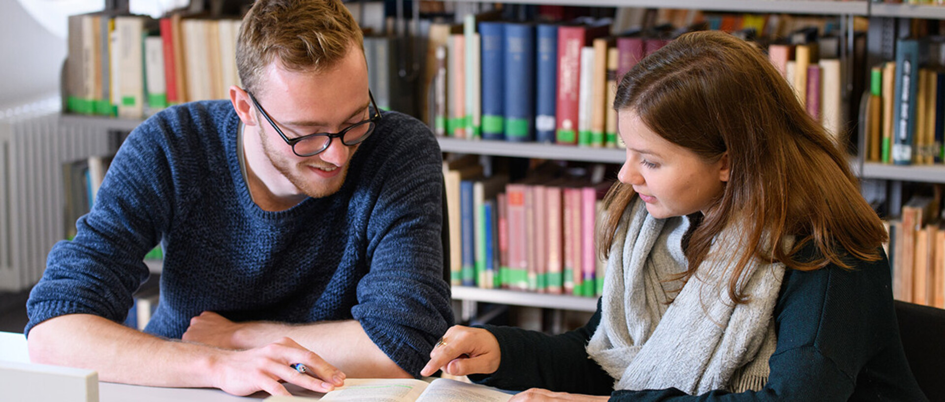 Eine Studentin und ein Student lesen gemeinsam in der Bibliothek ein Buch