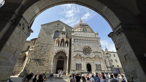 Blick durch einen Steinbogen auf die historische Piazza Vecchia in Bergamo mit der Fassade der Kirche Santa Maria Maggiore und vielen Besuchern