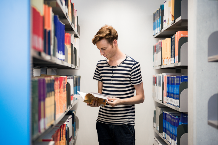 Student steht in einem Buch blätternd zwischen Bibliotheksregalen