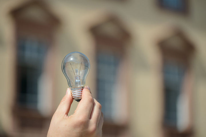 Eine Hand hält eine Glühbirne vor dem Mannheimer Schloss hoch.