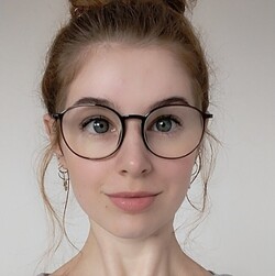 Porträt von Cora Beck. Cora Beck trägt einen grauen Hoodie und eine große runde Brille. Sie hat braune Augen und die braunen Haare zu einem Dutt nach oben gebunden.
