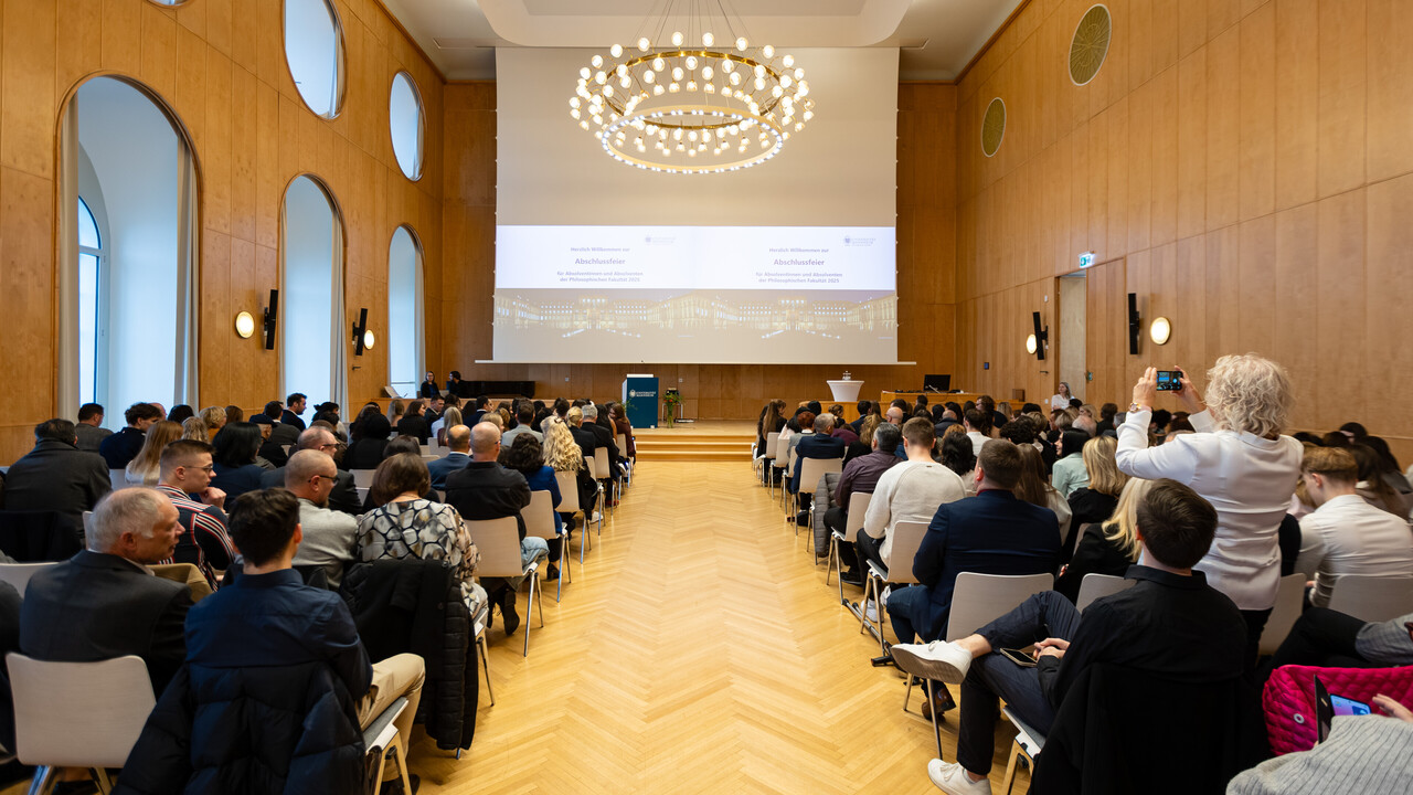 Blick von hinten in eine gut gefüllte Aula während einer Abschlussfeier. Das Publikum sitzt in Reihen und schaut auf die Bühne mit Präsentation.