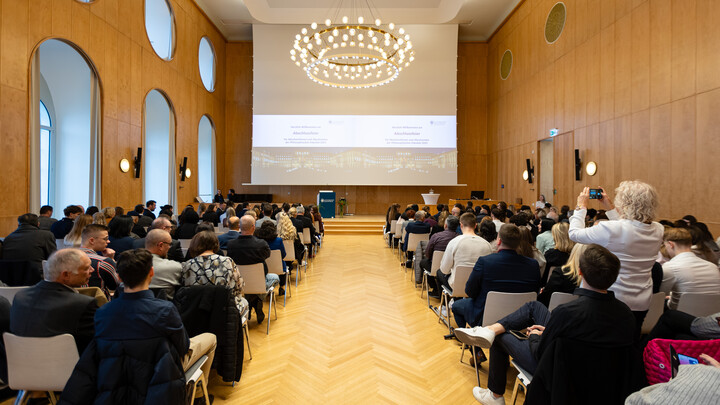 Blick von hinten in eine gut gefüllte Aula während einer Abschlussfeier. Das Publikum sitzt in Reihen und schaut auf die Bühne mit Präsentation.