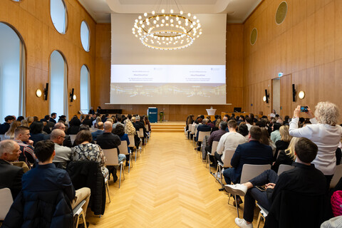 Blick von hinten in eine gut gefüllte Aula während einer Abschlussfeier. Das Publikum sitzt in Reihen und schaut auf die Bühne mit Präsentation.