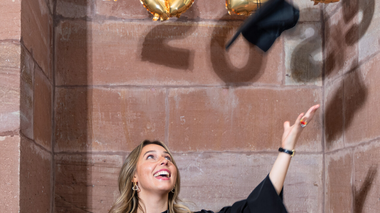 Eine Absolventin im Talar wirft freudig Ihren Hut in die Luft. Sie steht vor einer Wand, die mit goldenen Ballons im Schriftzug 2025 dekoriert ist.
